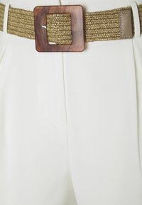 Wovený béžový opasek s obdélníkovou dřevěnou sponou, doplněný o světlé kalhoty z látky se záhyby a bočními kapsami.