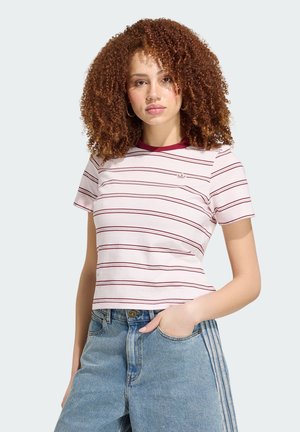 Jeune femme aux cheveux bouclés portant un t-shirt rayé blanc et bordeaux et un jean bleu taille haute, main dans la poche, sur fond uni.