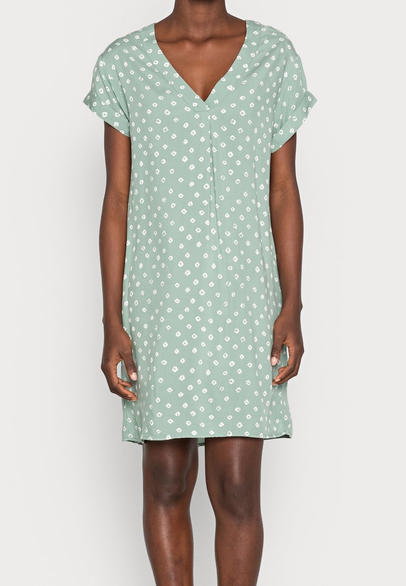 Femme portant une robe vert clair à manches courtes avec des motifs losanges blancs et un décolleté en V, debout devant un fond uni.