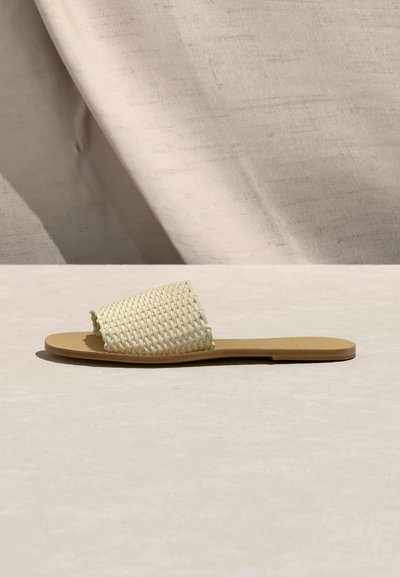 Επίπεδη σαγιονάρα με καφέ σόλα και υφασμάτινο, φαρδύ εκρού λουράκι, τοποθετημένη σε ανοιχτό μπεζ φόντο με υφή υφάσματος και επιφάνειας.