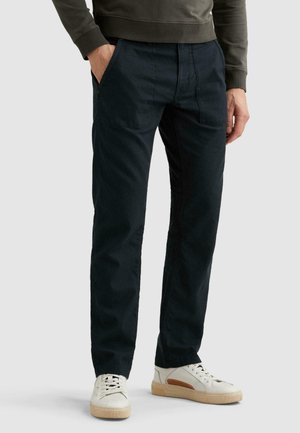 Muž nosi čierne nohavice s rovnými nohavicami a prednými našitými vreckami, v kombinácii s bielymi teniskami a tmavým tričkom s dlhým rukávom, stojaci na sivej podlahe.