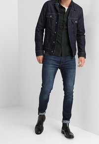 Dunkle Denimjacke mit zwei Vordertaschen, darüber ein dunkelgrünes Hemd mit Knöpfen. Schlanke blaue Jeans und schwarze Lederstiefel bis zum Knöchel.