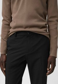 Suéter de punto marrón combinado con pantalones negros ajustados, con cierre de botón y bolsillos laterales. La tela parece suave y estructurada.