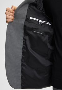 Grauer Blazer mit strukturierter, gewebter Außenseite und glattem schwarzem Innenfutter. Verfügt über dehnbaren Stoff und eine Innentasche mit Knopf.