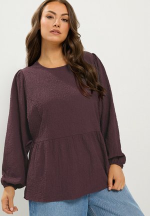 Vrouw die een gestructureerde donkerpaarse blouse met lange mouwen en lichtblauwe jeans draagt tegen een effen lichte achtergrond.