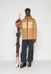 Vyras rusvame slidinėjimo džemperyje, juodose sniego kelnių, slidinėjimo batais ir akinių nuo saulės laikantis slidžių vertikaliai priešais vientisą foną.