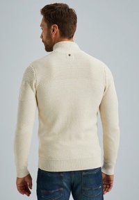 Strickpullover in Creme mit hohem Kragen, strukturiertem Muster, gerippten Bündchen und Saum, mit einem Etikett am linken Ärmel und einem Logo auf der Rückseite.