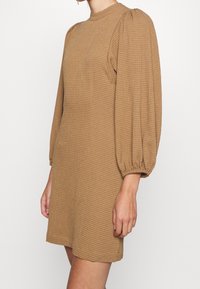 Mujer vistiendo un mini vestido de manga larga texturizado color beige, con mangas abullonadas y un cuello alto, de pie contra un fondo liso.
