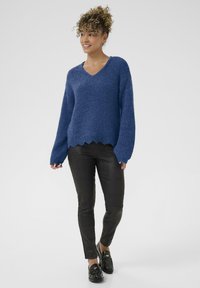 Blauer Strickpullover mit V-Ausschnitt und welligem Saum, kombiniert mit schwarzen Kunstlederhosen mit strukturiertem Muster. Schwarze Mokkasins runden den Look ab.