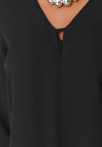 Närbild av en svart topp med en liten framklippning och ett silverpärlarmband mot brun hud.