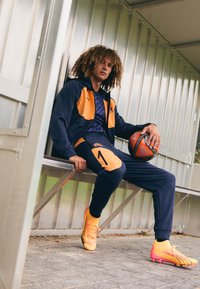 Sportoutfit bestaande uit een marineblauwe en oranje jack met capuchon, bijpassende broek en feloranje voetbalschoenen, met een voetbal in de hand.
