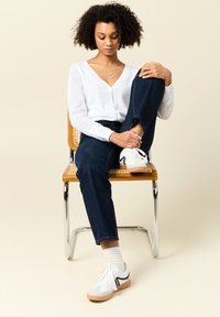 Vit knapp-up cardigan med nätärmar, mörkblå denimjeans och vita sneakers med svarta detaljer, sittande på en trästol.