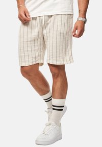 Weiße, strukturierte Shorts mit vertikalen schwarzen Streifen, ausgestattet mit einem elastischen Bund und Kordelzug. Kombiniert mit weißen Sneakers und gestreiften Socken.