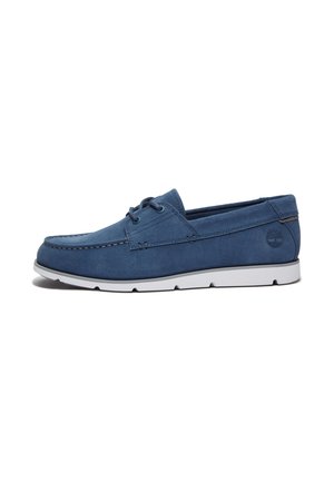 Chaussure bateau en daim bleu avec semelle en caoutchouc blanche et détails de lacets, vue latérale avec coutures visibles et logo de la marque près du talon.