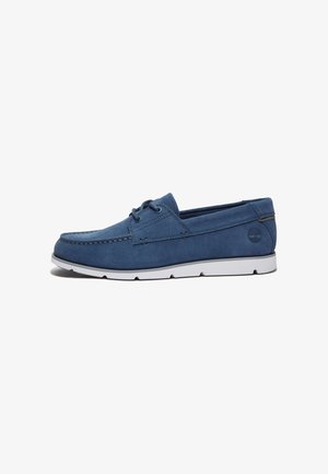 Chaussure bateau en daim bleu avec semelle en caoutchouc blanche et détails de lacets, vue latérale avec coutures visibles et logo de la marque près du talon.