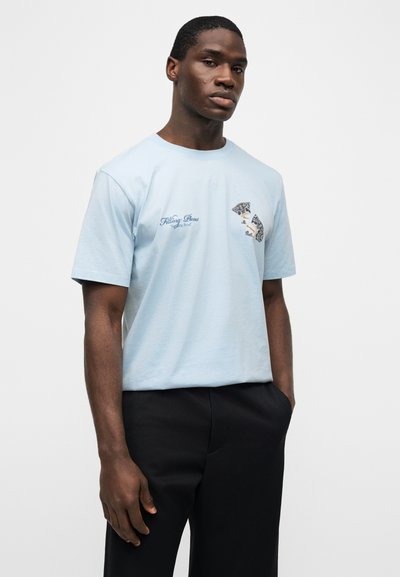 Άνδρας που φοράει γαλάζιο μπλουζάκι με γραφικό κείμενο και σχέδια στο στήθος, συνδυασμένο με μαύρο παντελόνι, στέκεται μπροστά σε λευκό φόντο.