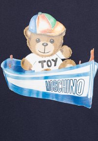 Námořnické tričko s potiskem kresleného medvěda, který nosí vícero barevnou čepici a bílé tričko s nápisem "TOY," sedícího v modré a bílé lodi s nápisem "MOSCHINO."