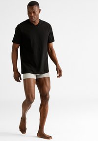 Zwarte v-hals t-shirt, ontspannen pasvorm, gecombineerd met lichtgrijze boxershorts. Model staat op blote voeten en toont huidskleur en lichaamsbouw.