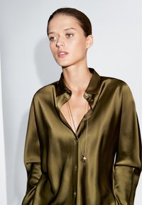 Olive green satin shirt with a collar, featuring a button-up design and a deep neckline. A long silver necklace with a round pendant is visible.