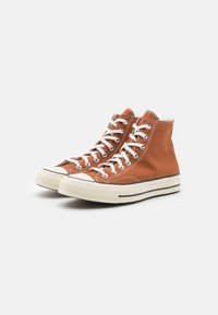 Sneaker alte in tela marrone, con lacci bianchi, punta in gomma e suola in gomma striata. Dettagli di cucitura distintivi sulla tomaia.