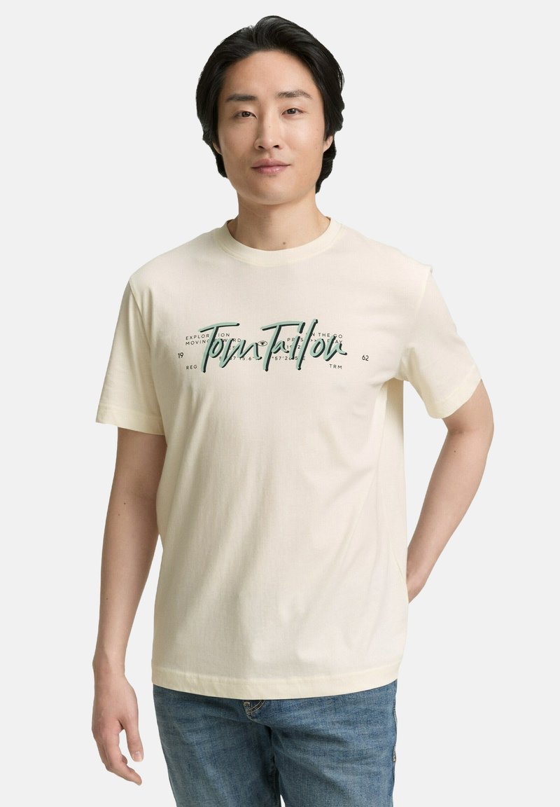 Licht crèmekleurig katoenen T-shirt met korte mouwen, met groene bedrukte tekst en ontwerpelementen aan de voorkant, gecombineerd met een blauwe spijkerbroek.