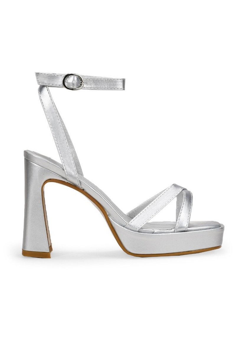 Sandalias plateadas de tacón alto con un acabado metálico brillante, tira en el tobillo y bandas cruzadas en los dedos, presentando un tacón grueso y plataforma.