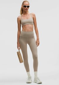 Sujetador deportivo beige y leggings de cintura alta hechos de una tela suave y elástica. La modelo sostiene una botella de agua marrón con la marca "lululemon".