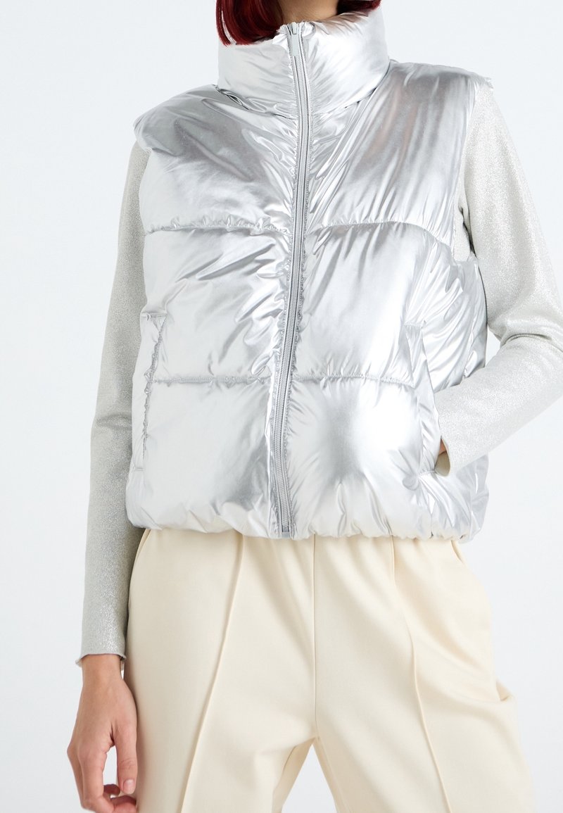 Silberne gesteppte Weste mit hohem Kragen und durchgehendem Reißverschluss. Verfügt über glänzende Textur, Seitentaschen und ein gesteppte Design; kombiniert mit einem leichten Pullover.