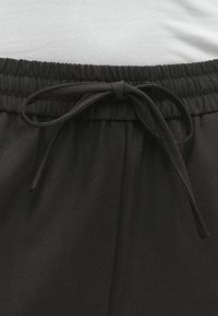 Melnas sporta bikses ar elastīgu jostasvietu, priekšā ar saites aizdari un mīkstu tekstūru. Tuvplāns, kas izceļ jostasvietas detaļas.