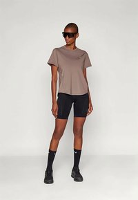 Pikk inimene, kellel on seljas üle suure t-särgi pruun, mustad jalgrattashorts, mustad Nike sokid, mustad spordijalatsid ja suured mustad prillid, seisab valge tausta ees.