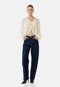 Cremefarvet bluse med flæseaccentuer, bundet foran, parret med mørkeblå høj talje jeans og sorte spidse hæle. Simpel, flydende design.
