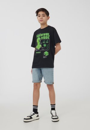 T-shirt di cotone nero con una grafica verde e il testo "SURVIVAL MODE". Abbinato a pantaloni shorts in denim arrotolati e sneakers in bianco e nero.