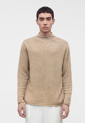 Giovane uomo con capelli neri corti che indossa un maglione beige a collo alto e pantaloni bianchi, in piedi davanti a uno sfondo chiaro e uniforme.