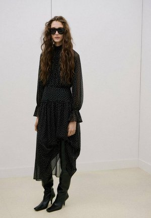 Mujer joven con cabello largo y rizado, vestida con un vestido negro semitransparente de lunares, botas negras hasta la rodilla y grandes gafas de sol negras, de pie en el interior.