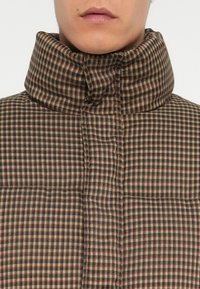 Gequilted jas met een hoge kraag in een bruin en crème ruitpatroon, met rode en zwarte accenten. Structuurstof met zichtbare stiksels.