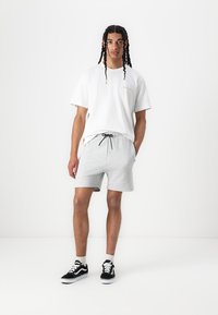 Camiseta blanca de algodón con cuello redondo, combinada con pantalones cortos grises que cuentan con una cinturilla elástica y cordón ajustable. Zapatillas negras.