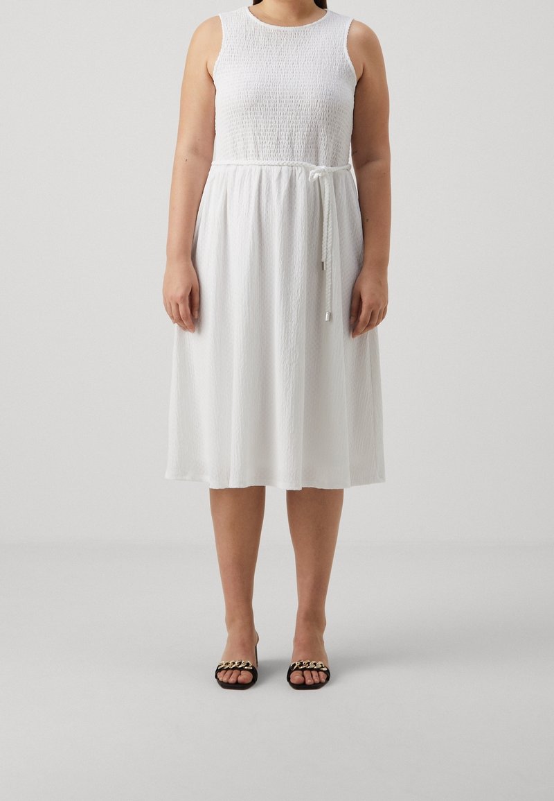 Robe blanche sans manches avec un corsage texturé et une jupe fluide, cintrée à la taille avec un lien. Associée à des sandales noires à lanières.