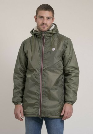 Homme portant une veste d'hiver à capuche vert olive avec fermeture éclair à l'avant et jean bleu clair, debout devant un fond uni.