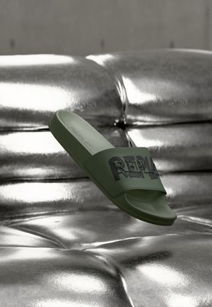 Sandale à glissière verte avec une bride texturée embossée du mot « REPLAY ». Semelle intérieure lisse et semelle en caoutchouc. Présentée sur un fond métallique brillant.