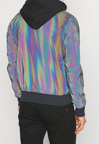 Irydescentna kurtka bomberka z kapturem, o gładkiej teksturze, elastycznymi mankietami, szarym prążkowanym pasem w talii oraz kontrastującym czarnym kapturem.