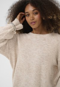 Bež džemper u velikom formatu izrađen od mekanog, teksturiranog materijala sa okruglim izrezom i rasparenim rukavima. Ima suptilan uzorak i rebraste manšete.