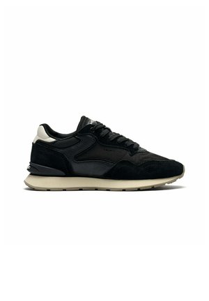 Zwart-witte lage sneaker met gestructureerde leren en suède bovenkant, zwarte veters en beige zool, zijkant gezien op een witte achtergrond.