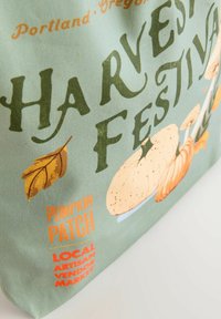 Borsa tote in tela con sfondo verde chiaro e la scritta "FESTIVAL DEL RACCOLTO" in verde scuro, illustrazioni di zucche e un'etichetta arancione "CAMPO DI ZUCCHINE".