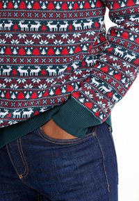 Adīta džemperis ar svētku rakstu, kurā attēloti ziemeļbrieži un koki sarkanos, baltos un zaļos toņos; ribotas piedurknes un apaļa kakla izgriezums.