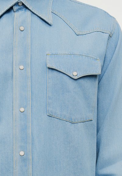 Φωτεινό γαλάζιο πουκάμισο από τζιν με κουμπιά πέρλες, αιχμηρό γιακά και μια μοναδική τσέπη στο στήθος με κίτρινες ραφές.