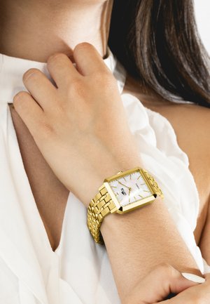 Zlatni metalni sat sa kvadratnim okvirom, bijelim brojčanikom i zlatnim oznakama sati; teksturirana, međusobno povezana narukvica; nosi se na zapešću svijetle puti.