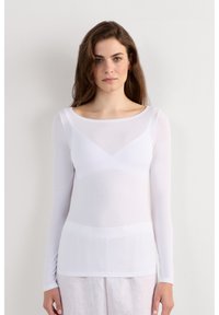 Jonge vrouw met lang bruin haar, gekleed in een doorschijnende witte lange mouwen top en lichtgekleurde broek, staand tegen een effen witte muur.