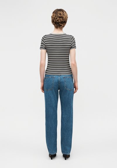 Άτομο με κοντά κυματιστά μαλλιά που φοράει μαύρο και άσπρο ριγέ μπλουζάκι, μπλε τζιν και μαύρα τακούνια, στέκεται γυρισμένο πλάτη σε απλό φόντο.