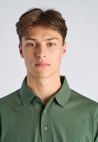 Jonge man met lichtbruin haar en hazelnootkleurige ogen, gekleed in een groen overhemd met kraag, kijkt recht vooruit tegen een effen lichte achtergrond.