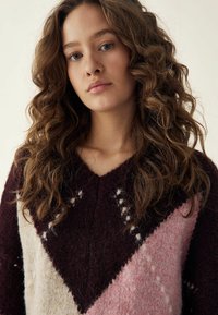 Pleteni pulover s V-izrezom, izrađen od guste, teksturirane tkanine; boje uključuju burgundac, krem i pastelno rozu s geometrijskim uzorcima.